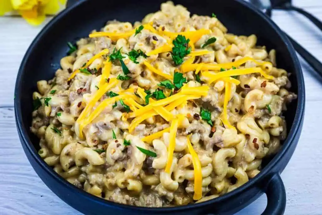 A black bowl filled with macaroni and ground beef in a creamy sauce, topped with shredded cheddar cheese and chopped parsley.