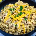 A black bowl filled with macaroni and ground beef in a creamy sauce, topped with shredded cheddar cheese and chopped parsley.