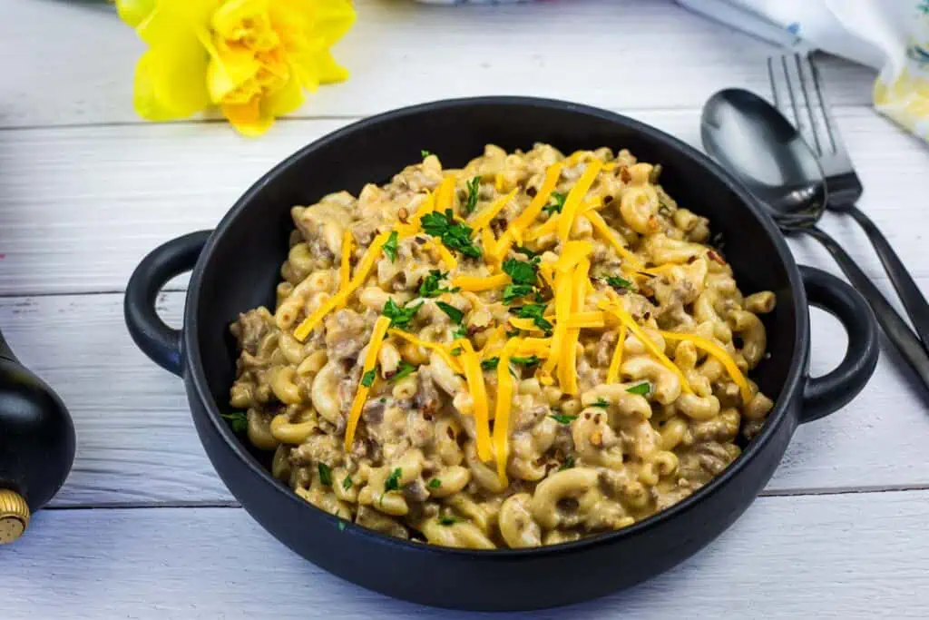 A black bowl filled with macaroni and ground beef in a creamy sauce, topped with shredded cheddar cheese and chopped parsley, placed on a white wooden surface.