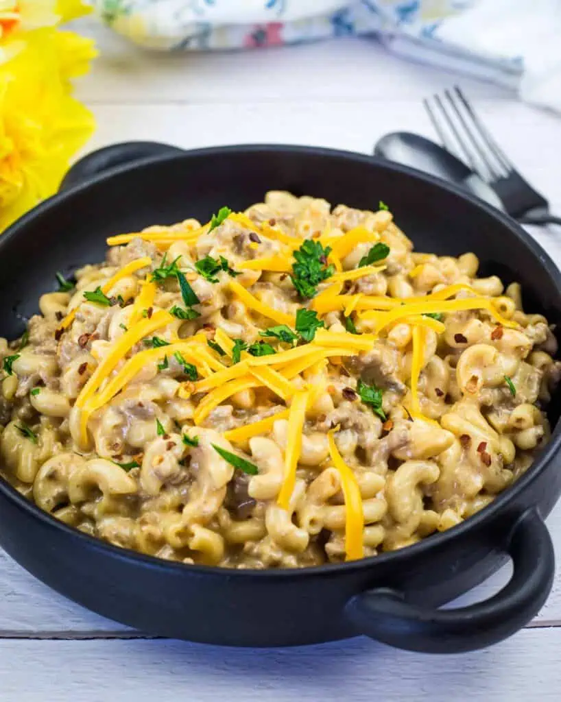 A black bowl filled with creamy macaroni and beef, topped with shredded cheese and chopped parsley, sits on a white wooden table with a fork and spoon nearby.