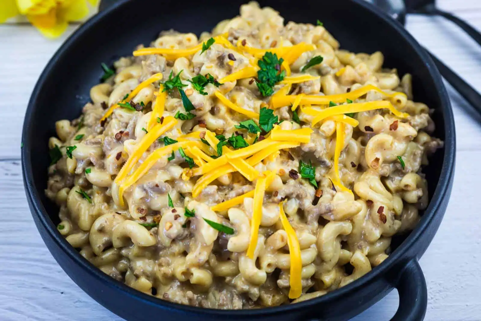 A black bowl filled with macaroni and cheese mixed with ground beef, topped with shredded cheddar cheese and chopped parsley.