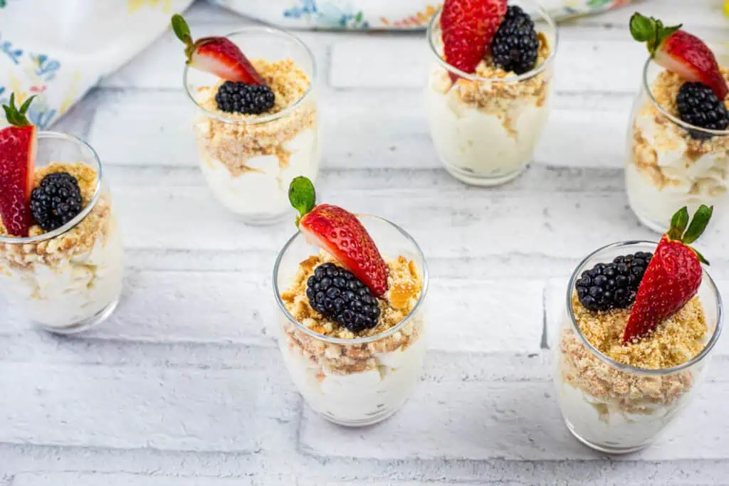 Six small glass cups filled with creamy dessert, topped with crushed crumbs, blackberries, and halved strawberries, arranged on a white surface.