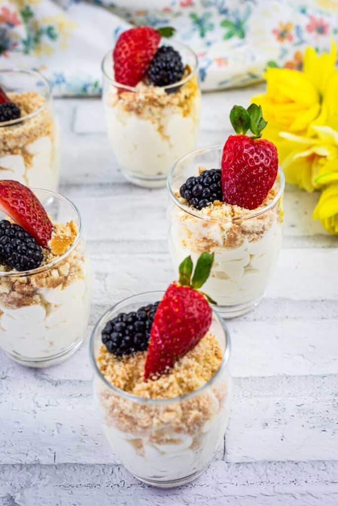 Five glass cups of layered dessert with whipped cream, crumbled topping, strawberries, and blackberries are arranged on a white wooden surface next to a yellow flower.