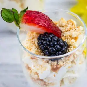 A dessert in a glass topped with a halved strawberry, a blackberry, and crumbled cookie or biscuit, with creamy layers beneath.
