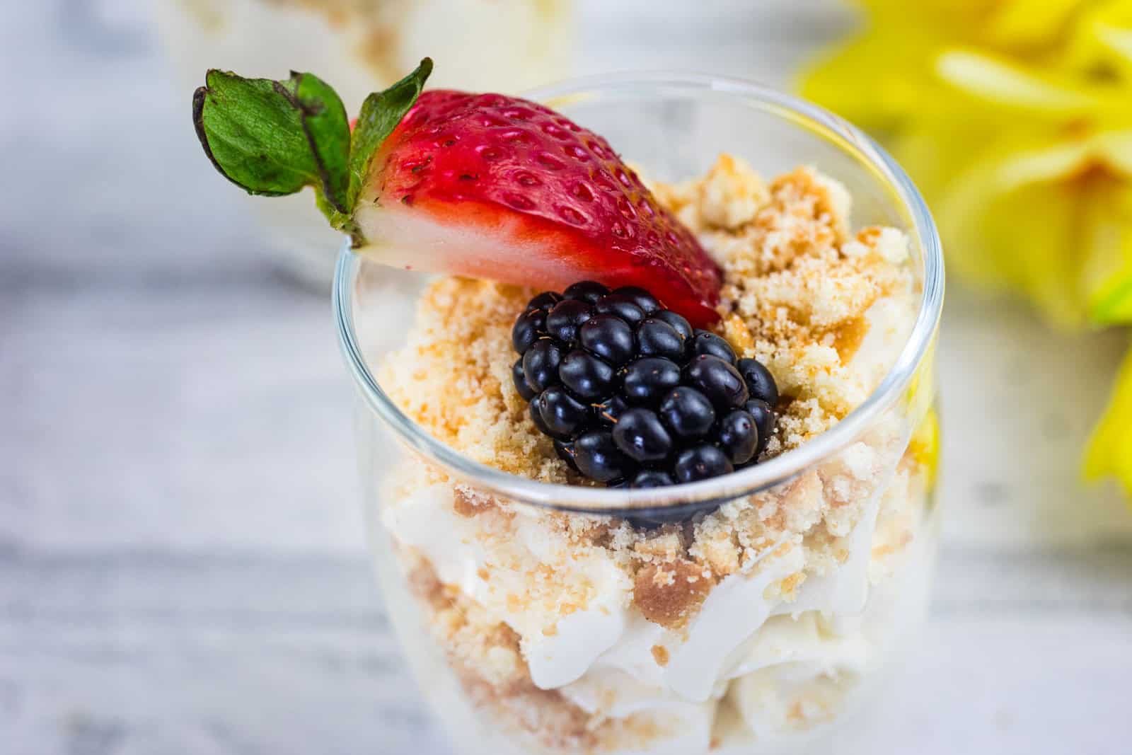 A dessert in a glass topped with a halved strawberry, a blackberry, and crumbled cookie or biscuit, with creamy layers beneath.