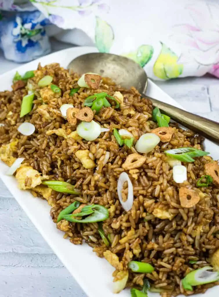A rectangular white plate with fried rice garnished with sliced green onions and crispy garlic, served with a metal spoon on a light wooden surface.