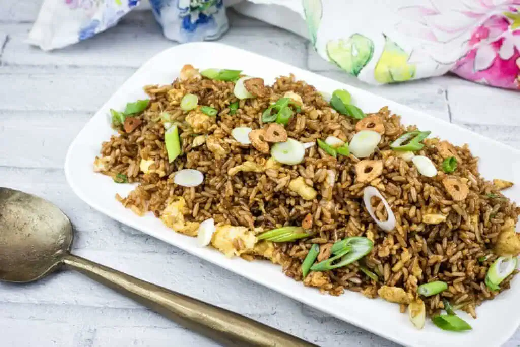 A rectangular white plate with fried rice garnished with sliced garlic and green onions, placed on a white wooden surface next to a brass spoon and floral napkins.