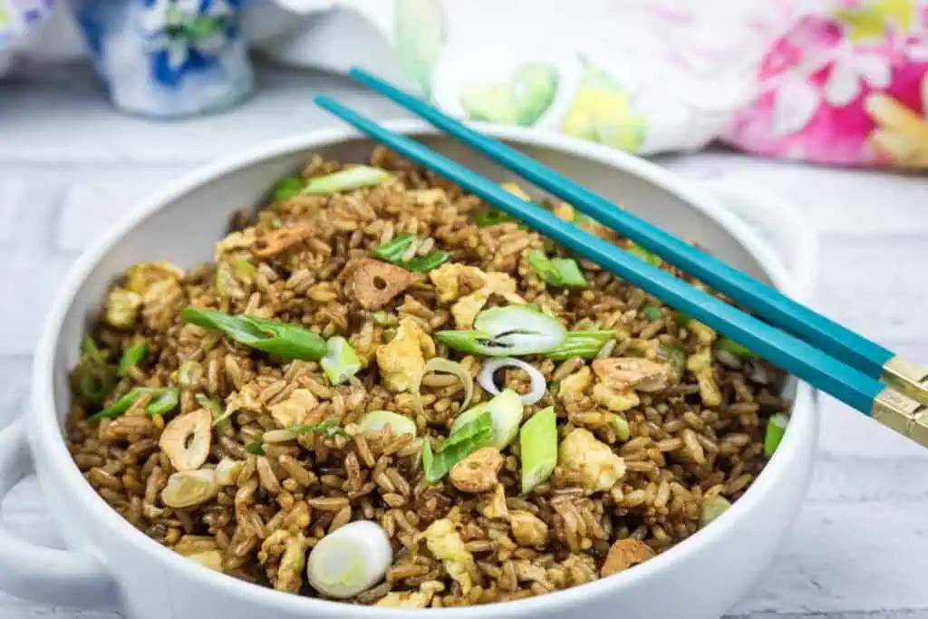 A bowl of fried rice with scrambled egg, sliced scallions, and crispy garlic, served with teal chopsticks on the side.
