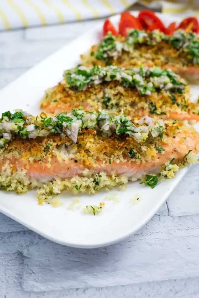 Three baked salmon fillets topped with a breadcrumb herb mixture and diced onions, served on a white plate with sliced tomatoes in the background.