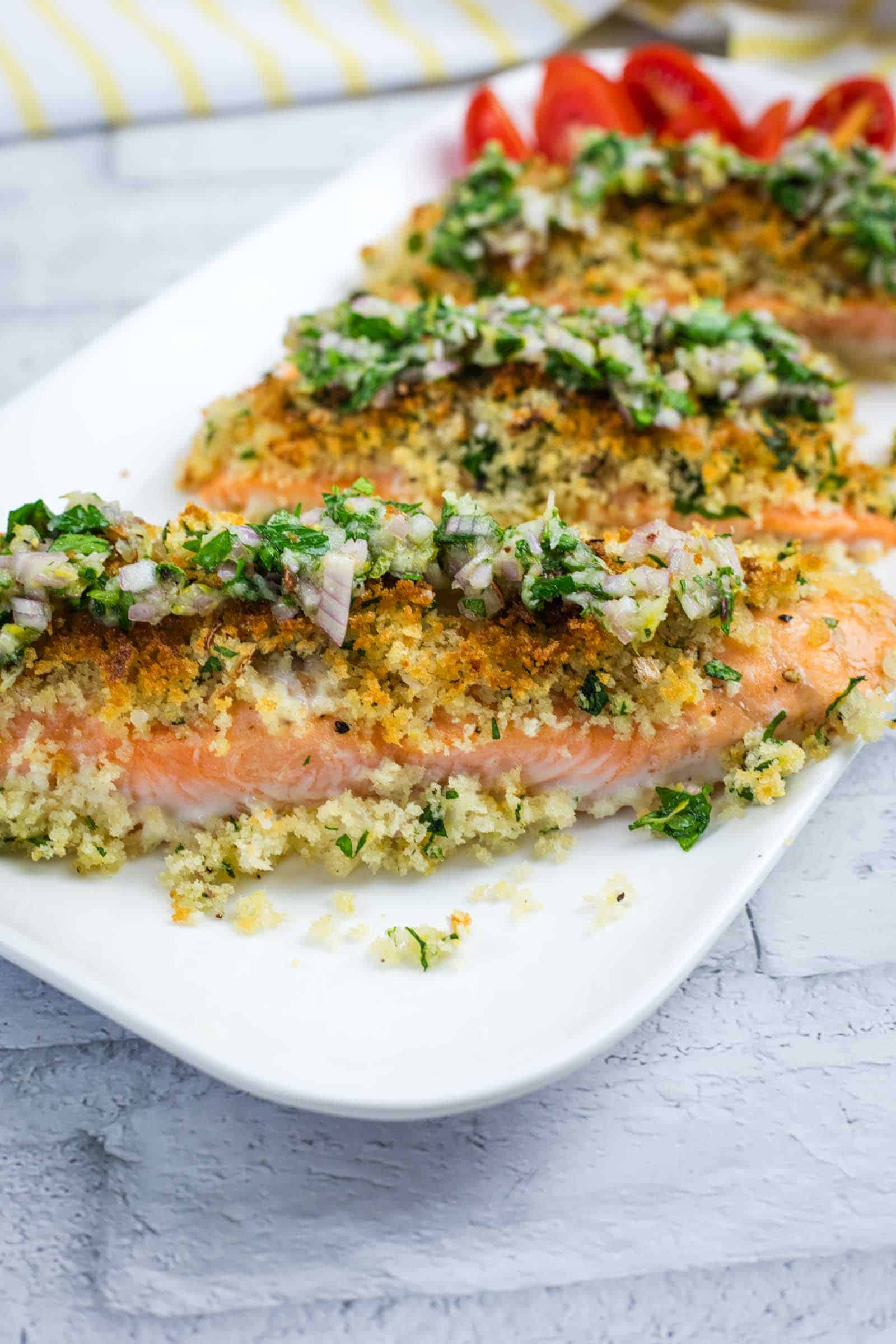 Three baked salmon fillets topped with a breadcrumb herb mixture and diced onions, served on a white plate with sliced tomatoes in the background.