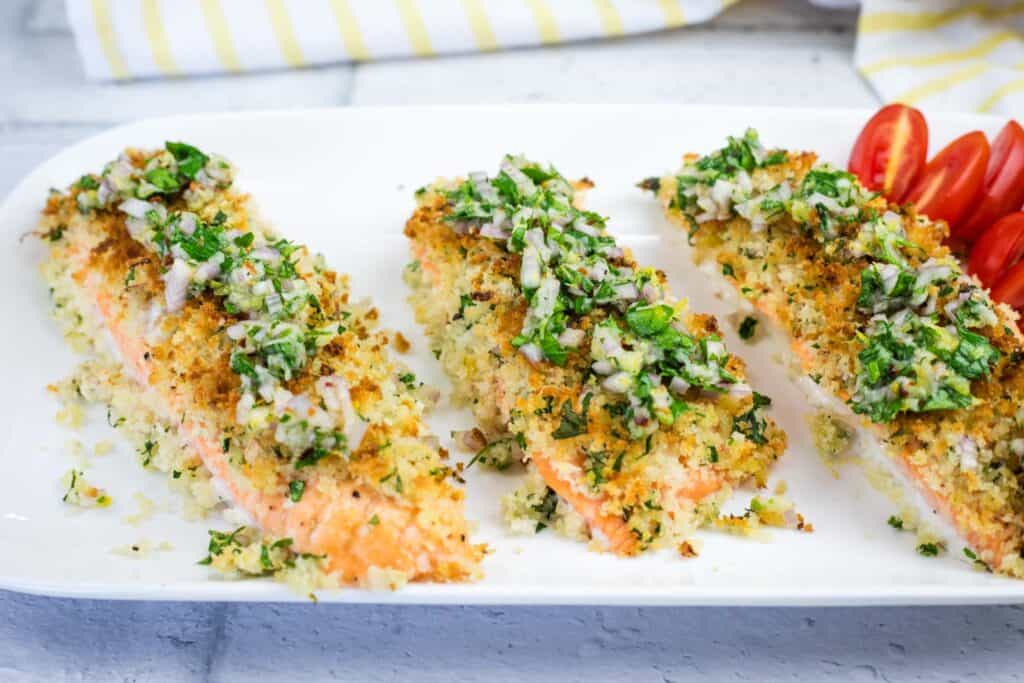 Three pieces of baked salmon topped with a herb and breadcrumb crust, served on a white plate with a side of sliced cherry tomatoes.