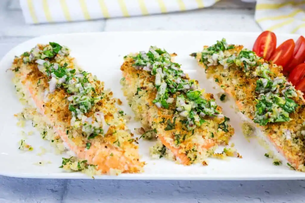 Three pieces of baked salmon topped with a herb and breadcrumb crust, served on a white plate with a side of sliced cherry tomatoes.