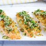 Three pieces of baked salmon topped with a herb and breadcrumb crust, served on a white plate with a side of sliced cherry tomatoes.