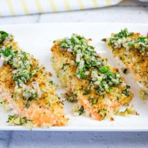Three pieces of baked salmon topped with a herb and breadcrumb crust, served on a white plate with a side of sliced cherry tomatoes.