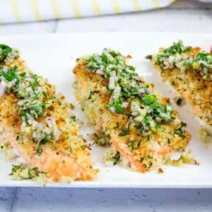 Three pieces of baked salmon topped with a herb and breadcrumb crust, served on a white plate with a side of sliced cherry tomatoes.