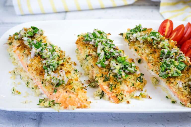 Three pieces of baked salmon topped with a herb and breadcrumb crust, served on a white plate with a side of sliced cherry tomatoes.