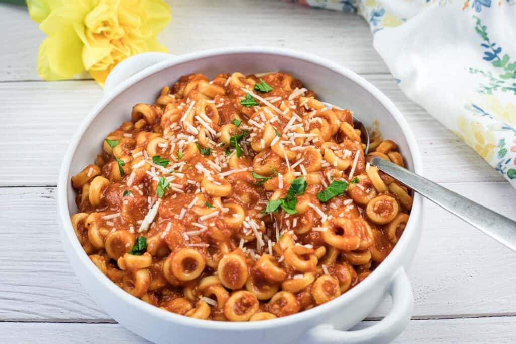 A white bowl filled with pasta in tomato sauce, garnished with grated cheese and chopped herbs, with a spoon inside. A yellow flower and floral cloth are in the background.