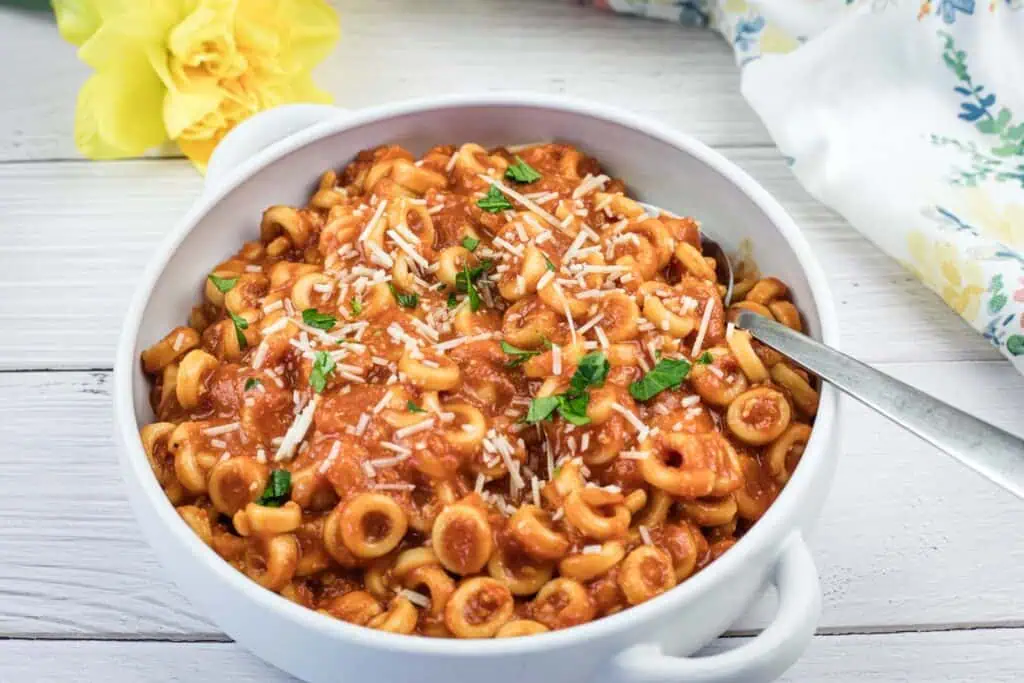 A white bowl filled with pasta in tomato sauce, garnished with grated cheese and chopped herbs, with a spoon inside. A yellow flower and floral cloth are in the background.