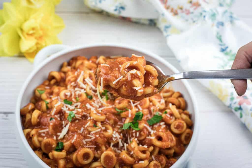 A bowl of pasta in tomato sauce topped with grated cheese and basil; a hand holds a forkful of pasta above the bowl.