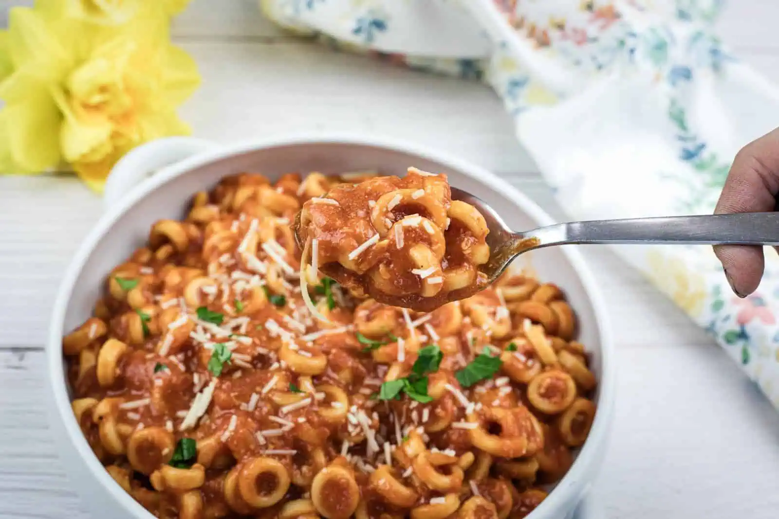A bowl of pasta in tomato sauce, topped with grated chaeese and chopped herbs, with a spoon lifting a portion; a yellow flower and a floral napkin are nearby.