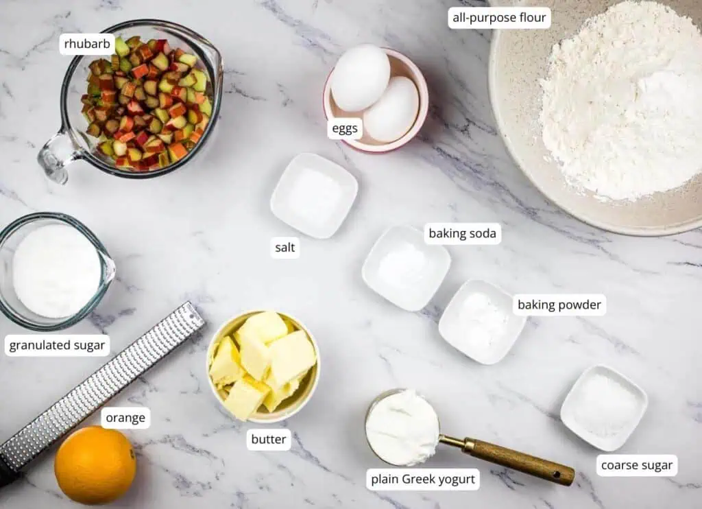A marble countertop displays labeled ingredients for baking, including rhubarb, eggs, flour, sugar, butter, yogurt, orange, salt, baking soda, baking powder, and coarse sugar.