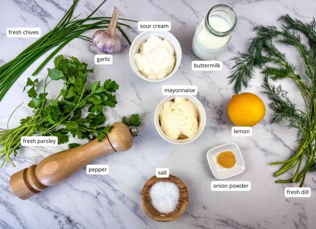 Ingredients for a dressing are arranged on a marble surface, including parsley, chives, dill, garlic, sour cream, mayonnaise, buttermilk, lemon, pepper, salt, and onion powder.