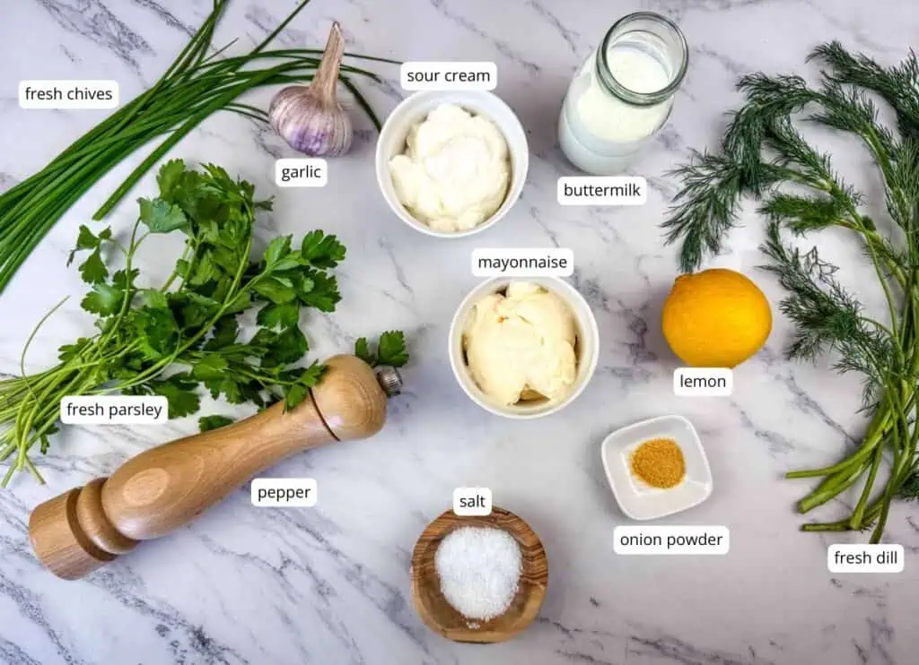Ingredients for a dressing are arranged on a marble surface, including parsley, chives, dill, garlic, sour cream, mayonnaise, buttermilk, lemon, pepper, salt, and onion powder.