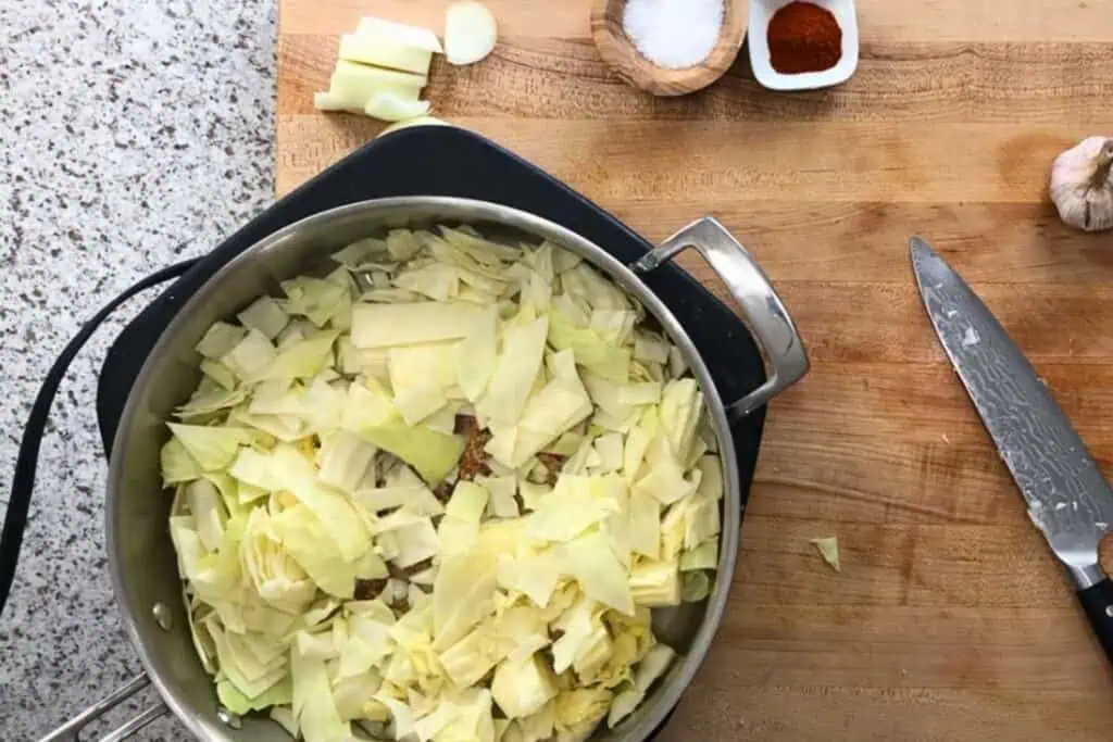 A saucepan filled with chopped cabbage sits on a wooden cutting board next to a knife, garlic, sliced ginger, salt, and spices.