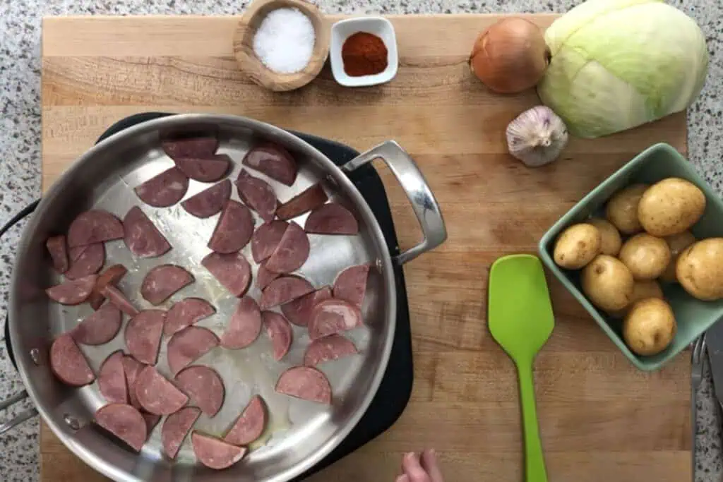 A skillet with sliced sausage on a stovetop, next to a cutting board holding potatoes, cabbage, onion, garlic, salt, and paprika.