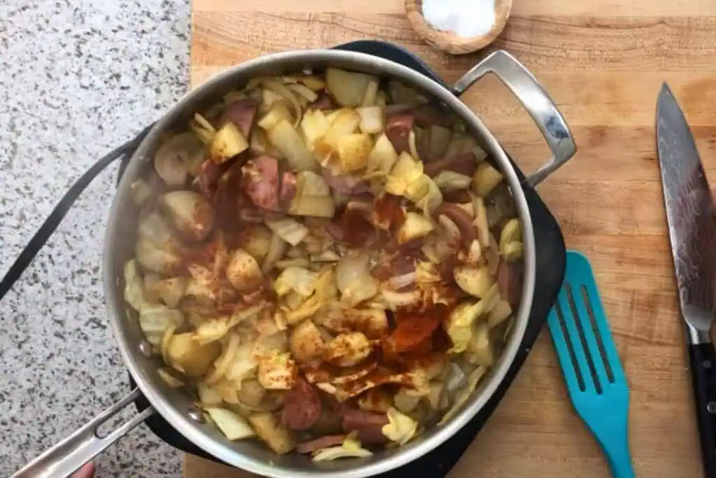A pot on a wooden cutting board contains chopped potatoes and onions being cooked with spices. A knife, pinch bowl of salt, and teal spatula are nearby.