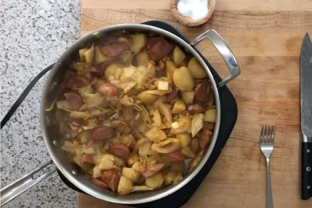 A pan filled with cooked potatoes, cabbage, and sausage sits on a wooden cutting board next to a fork, knife, and a small wooden bowl of salt.