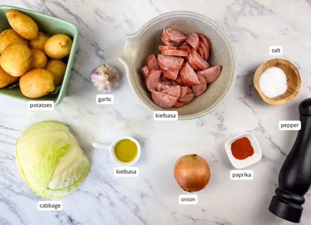 Ingredients arranged on a marble surface: potatoes, garlic, sliced kielbasa, cabbage, olive oil, onion, salt, pepper, and paprika, each labeled.