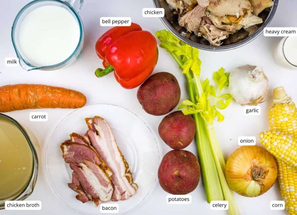 Assorted ingredients including milk, chicken broth, bacon, carrot, bell pepper, celery, potatoes, chicken, heavy cream, garlic, onion, and corn arranged on a white surface.
