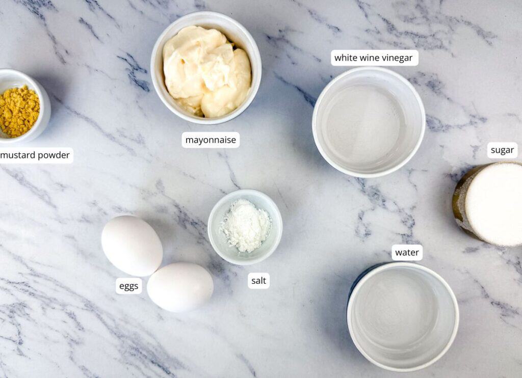 Ingredients on a marble surface: mustard powder, mayonnaise, white wine vinegar, sugar, water, salt, and two eggs, each labeled.
