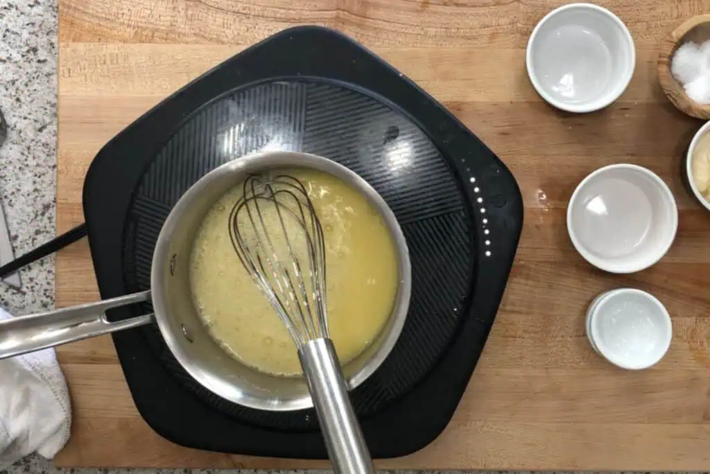 A saucepan with a whisk mixing a light yellow liquid sits on an electric cooktop, surrounded by small empty bowls on a wooden surface.