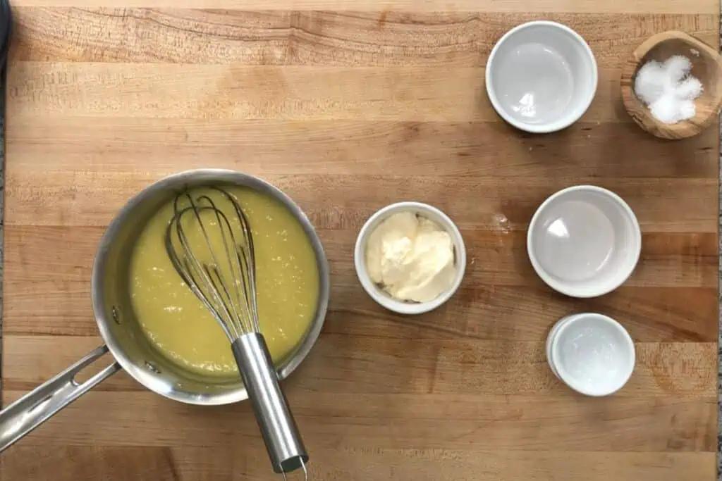 A saucepan with yellow sauce and a whisk, surrounded by small bowls containing mayonnaise, salt, and other ingredients on a wooden countertop.