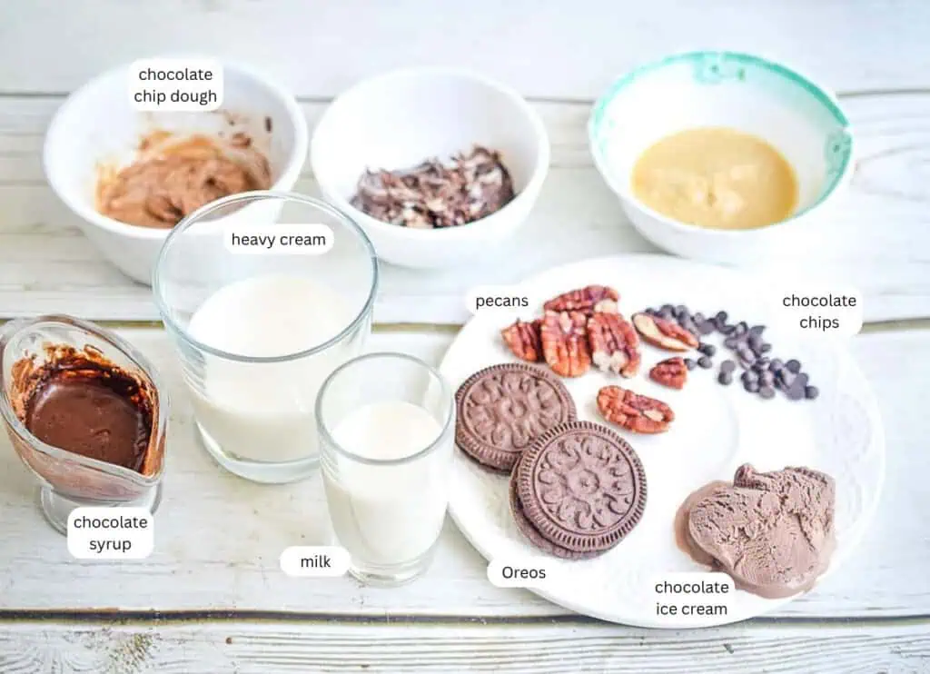 Various dessert ingredients, including milk, heavy cream, pecans, Oreos, chocolate chips, chocolate chip dough, chocolate syrup, and chocolate ice cream, are arranged on a white table.
