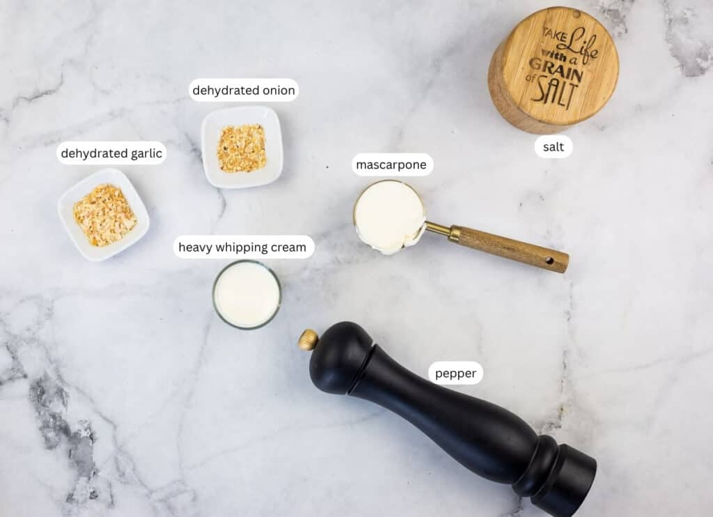 Ingredients on a white surface: bowls of dehydrated garlic and onion, a cup of heavy whipping cream, mascarpone in a measuring cup, salt container, and a pepper grinder.