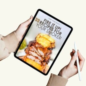 Person holding a tablet displaying a digital cookbook titled "Fire It Up! 33 Recipes for Your Smoker" with an image of stacked smoked meat and pineapple on the cover.