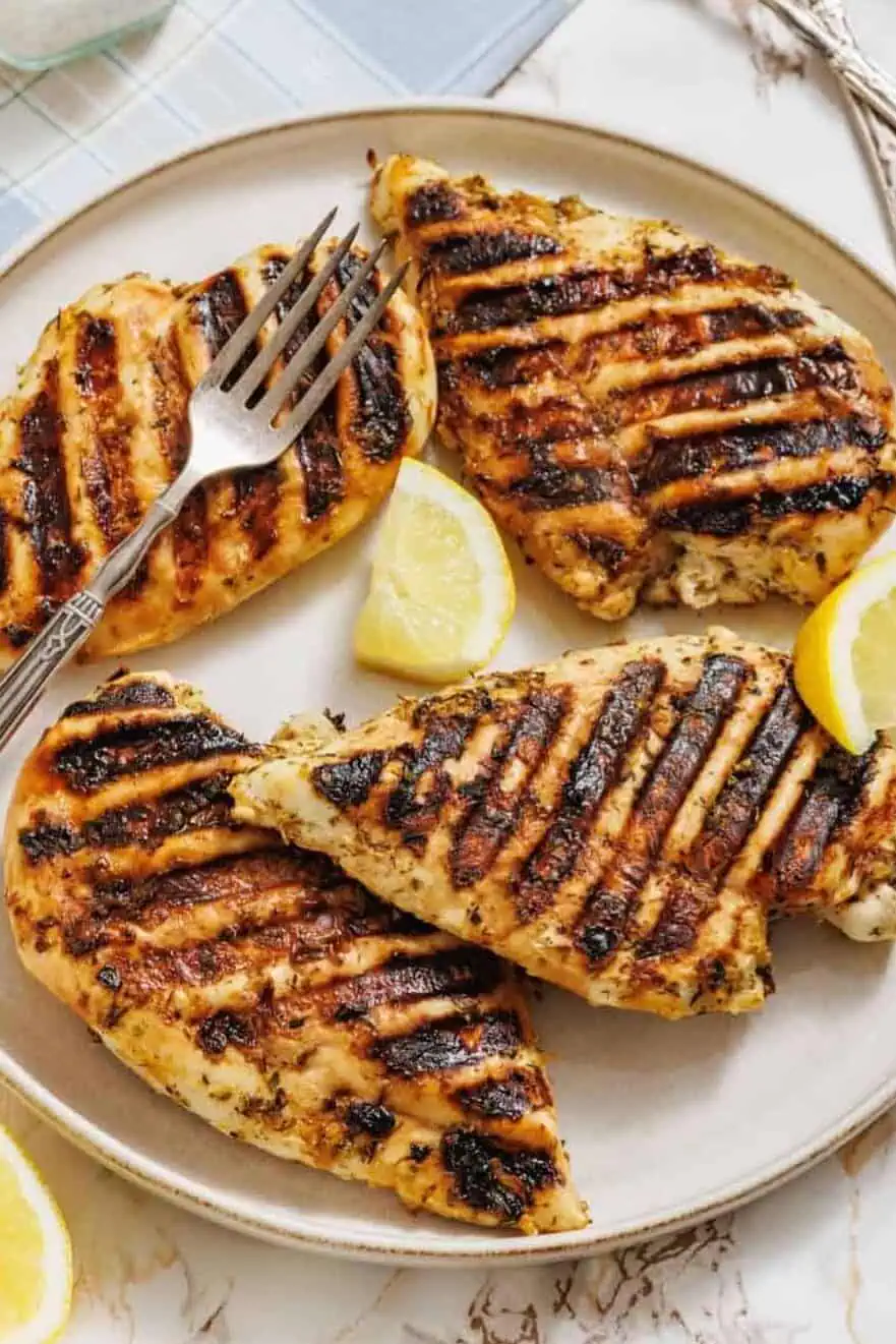 A plate with four pieces of grilled chicken breast, grill marks visible, garnished with lemon wedges; two forks, a jar of salt, and halved lemons are nearby on a table.