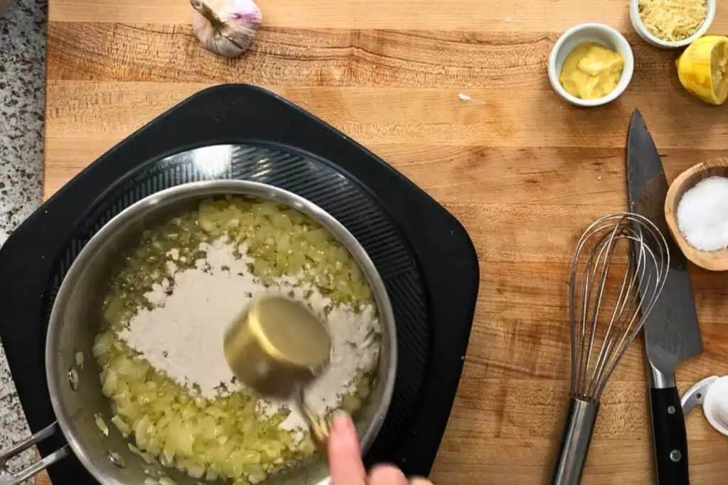 A hand adds flour to a pan of sautéed onions on an electric cooktop, with garlic, mustard, lemon, salt, and kitchen utensils on a wooden countertop nearby.