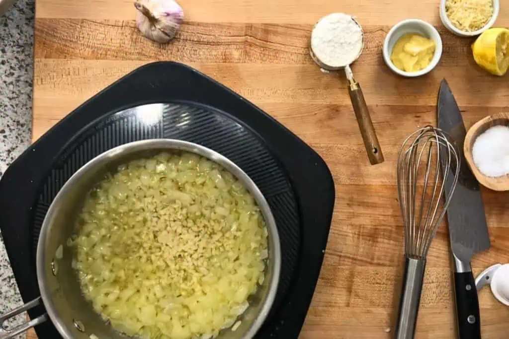 Overhead view of chopped onions and garlic sautéing in a pan on a wooden counter with ingredients, utensils, and a garlic bulb nearby.