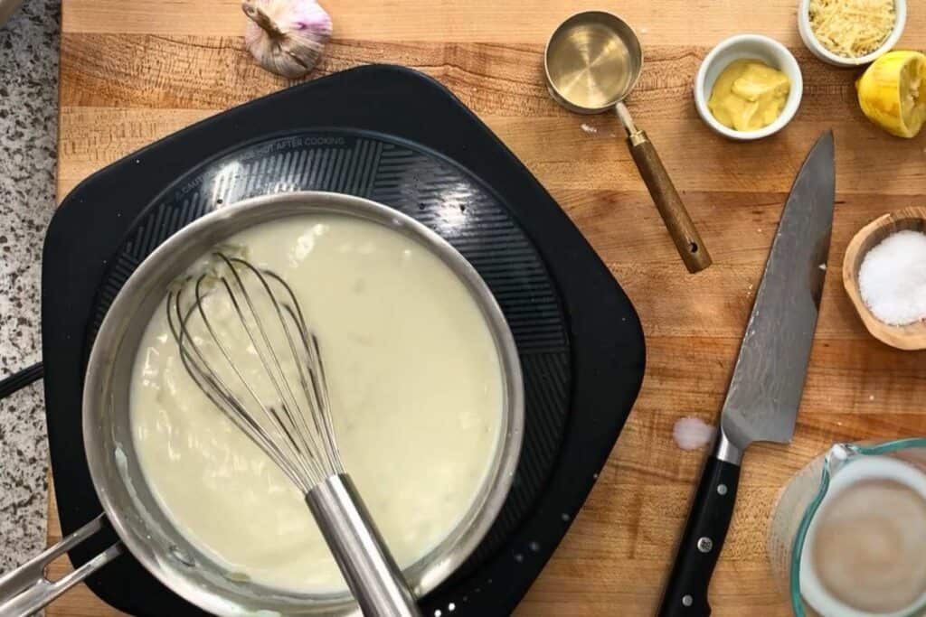 A whisk sits in a saucepan of white sauce on an electric cooktop beside a chef's knife, measuring cup, salt, mustard, lemon, and garlic on a wooden countertop.
