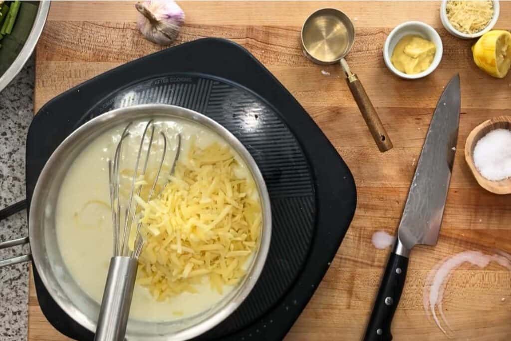 A metal pot with shredded cheese and a whisk sits on a stovetop, surrounded by a knife, measuring cup, garlic, mustard, lemon, and a bowl of salt on a wooden countertop.