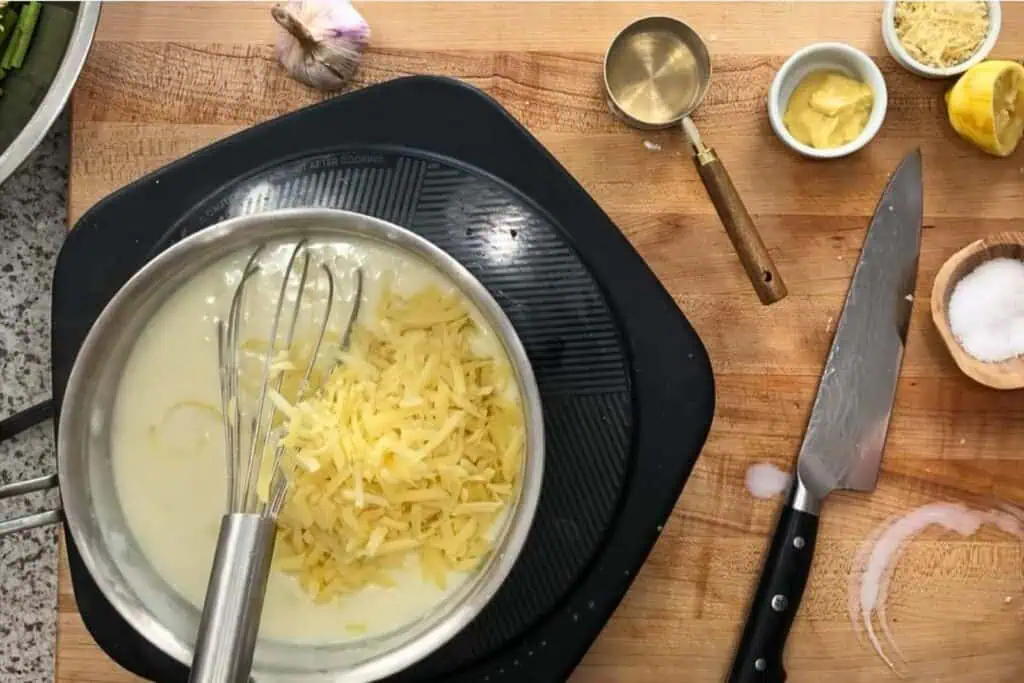 A metal pot with shredded cheese and a whisk sits on a stovetop, surrounded by a knife, measuring cup, garlic, mustard, lemon, and a bowl of salt on a wooden countertop.