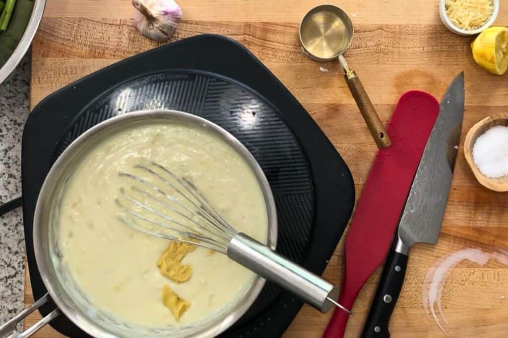 A saucepan with creamy sauce and a whisk sits on a stovetop; mustard, a chef's knife, measuring cup, salt, garlic, and lemon are on the wooden counter nearby.