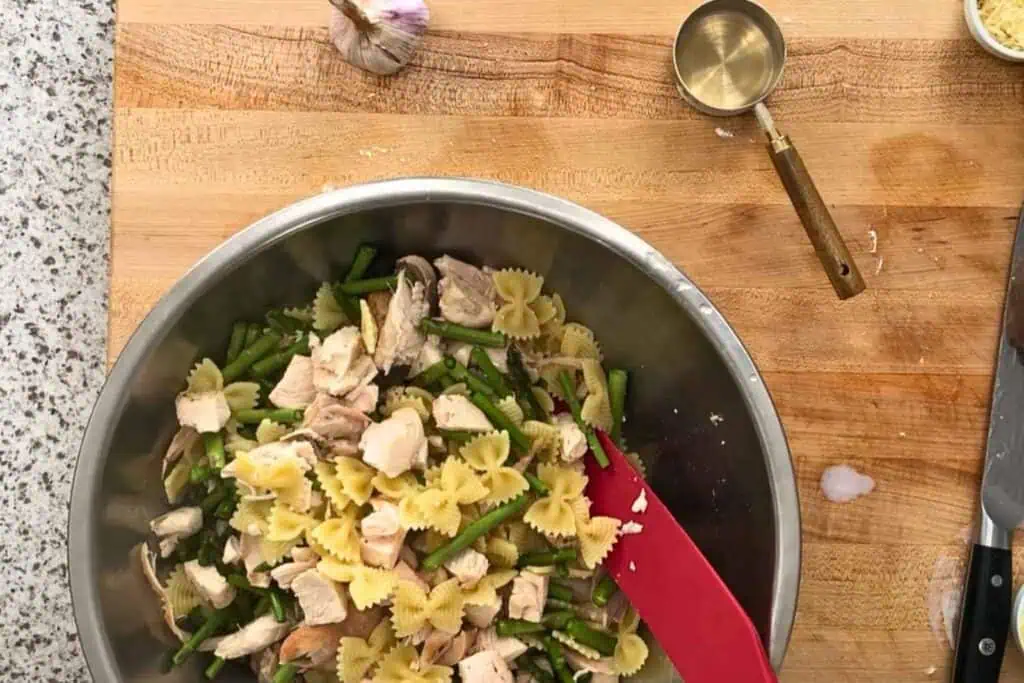 A stainless steel bowl with pasta, green beans, and chunks of chicken is on a wooden countertop with a red spatula, a measuring cup, garlic, and a knife nearby.