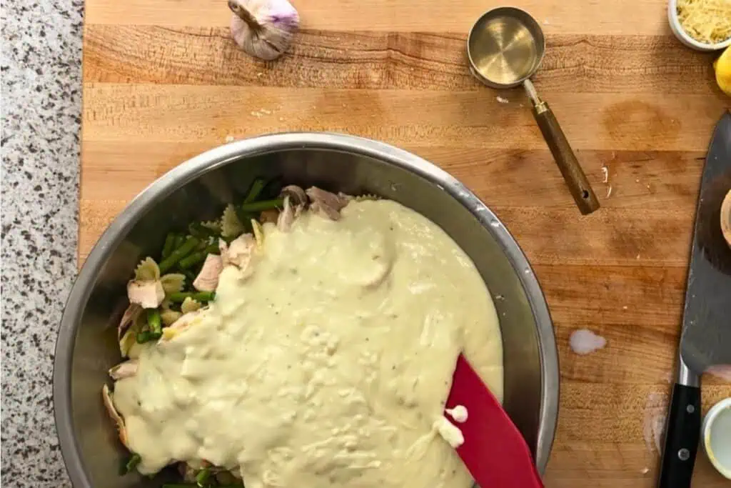 A mixing bowl of pasta and green beans topped with creamy sauce is being stirred with a red spatula on a wooden countertop. Measuring cup, garlic, and lemon are nearby.
