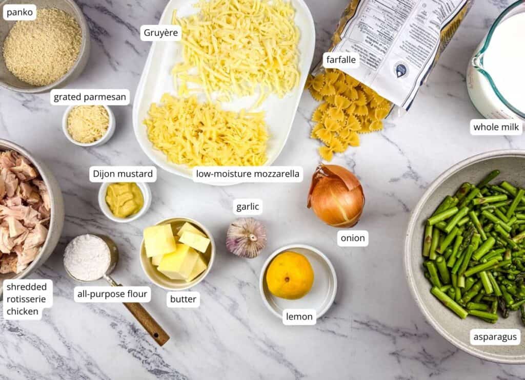 Labeled ingredients on a marble surface include farfalle, whole milk, Gruy&egrave;re, mozzarella, parmesan, panko, rotisserie chicken, flour, butter, Dijon mustard, garlic, onion, lemon, and asparagus.