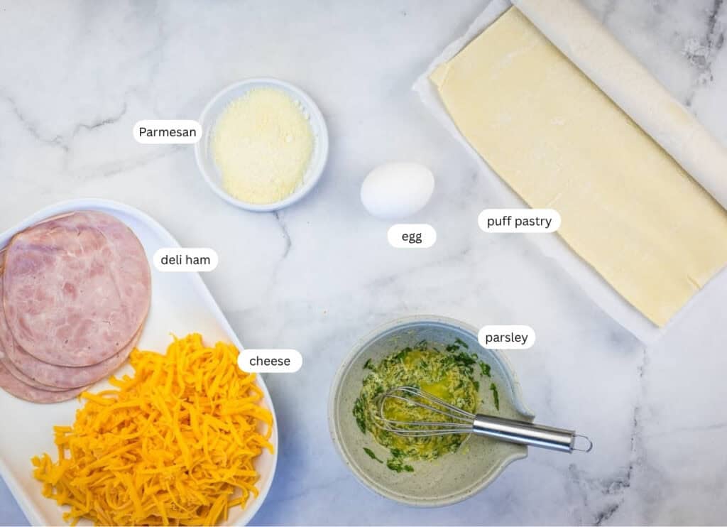 Sliced deli ham, shredded cheese, grated Parmesan, an egg, puff pastry sheet, and a bowl of whisked egg with parsley are arranged on a marble surface.